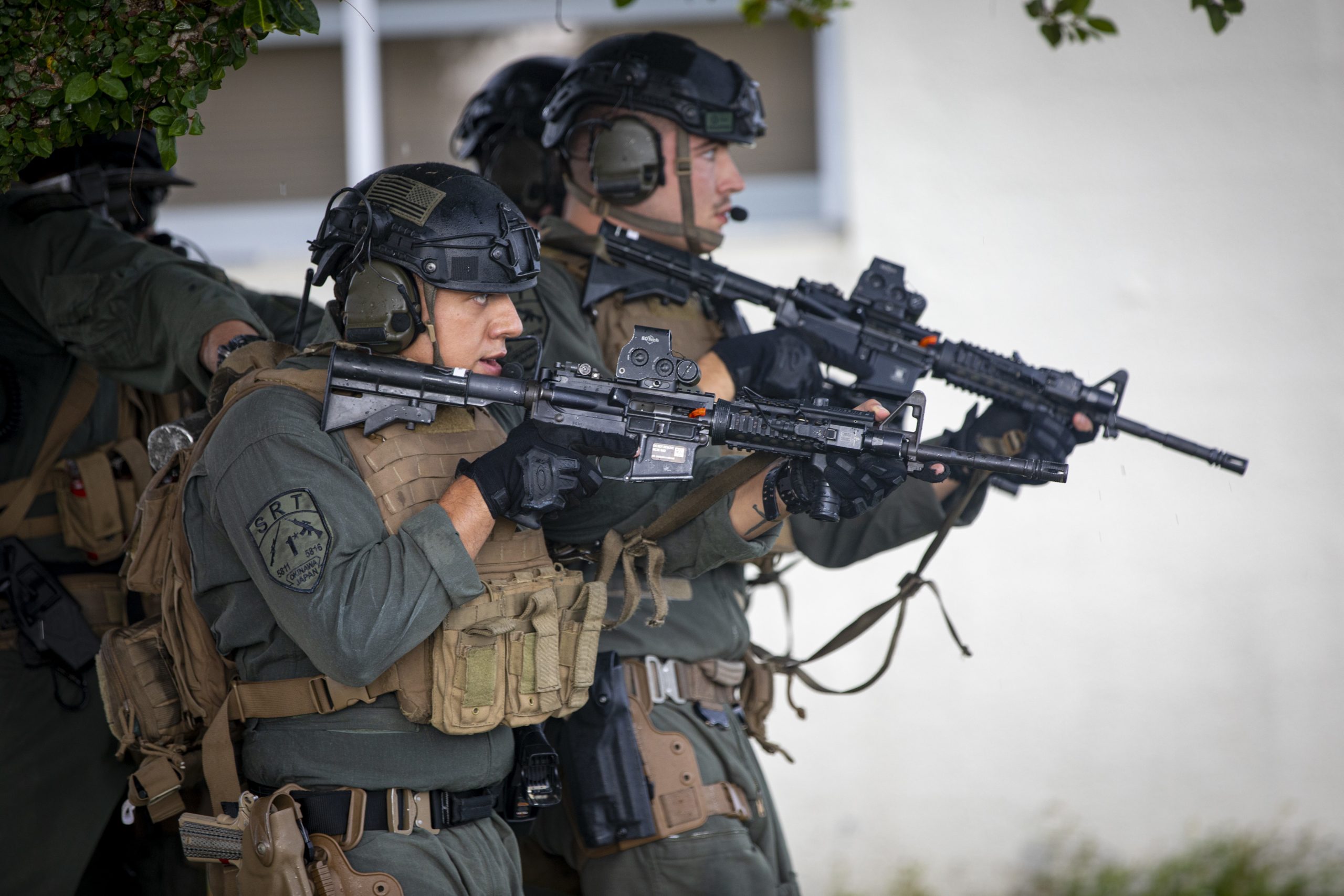 Three armed police officers in tactical gear and helmets aim rifles while standing in a defensive position near a building, partially under tree cover.